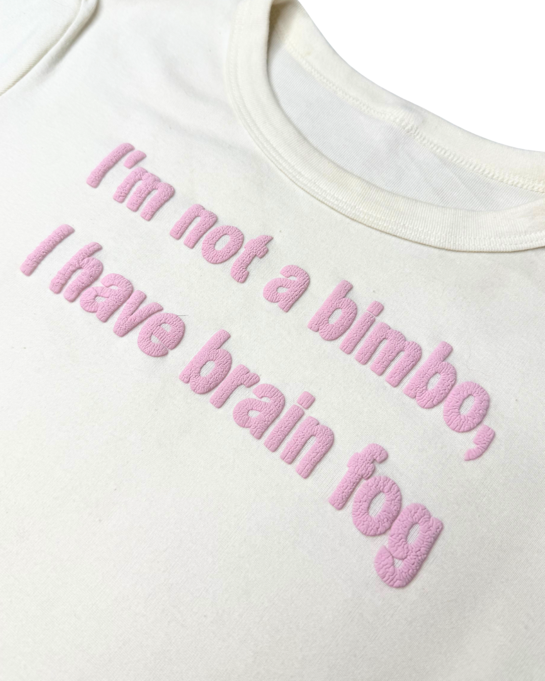 A close-up of an off-white crop top with a baby pink wrinkled puff vinyl design in the center that reads "I'm not a bimbo, I have brain fog". The wrinkled text resembled a brain. The background is white.