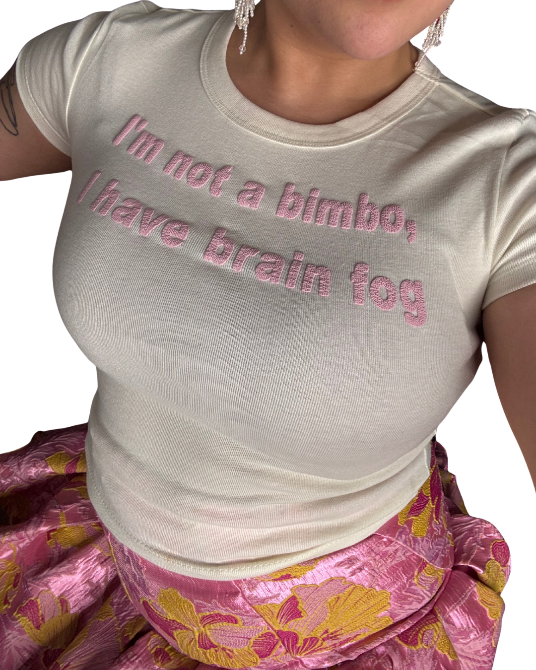 An off-white crop top with a baby pink wrinkled puff vinyl design in the center that reads "I'm not a bimbo, I have brain fog". The wrinkled text resembled a brain. The background is white. A woman is wearing he shirt with a pink skirt.